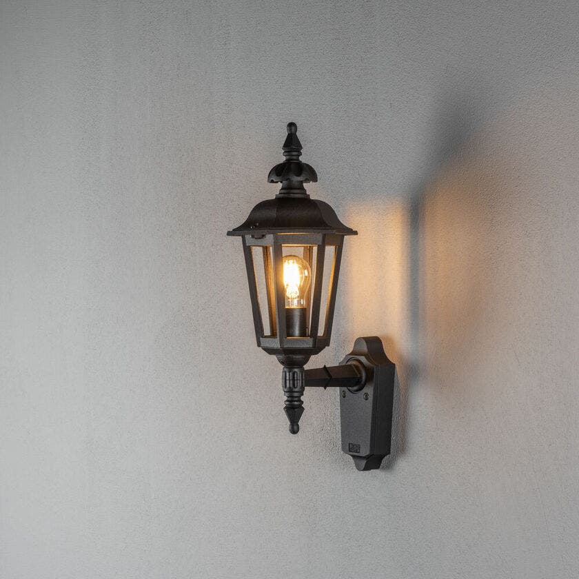 Konstsmide Pallas 518-750 svart utomhusvägglykta monterad på en grå fasad med en tänd E27-glödlampa
